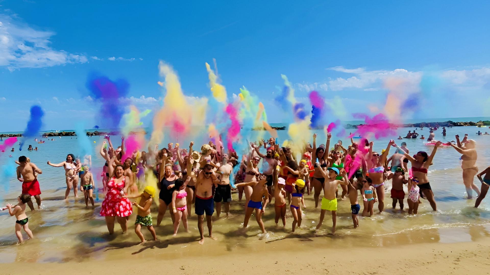 Persone festeggiano sulla spiaggia lanciando polveri colorate nell'aria.