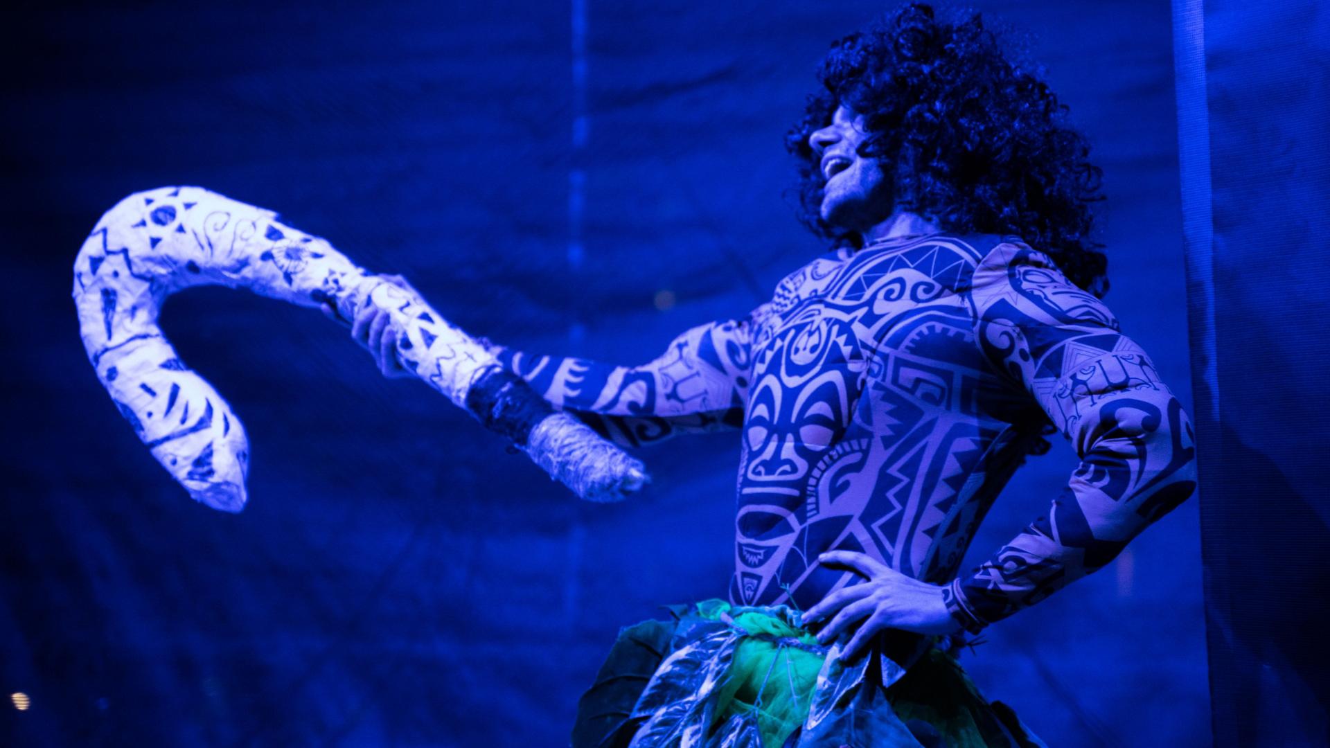 Uomo in costume tribale con tatuaggi e un grande bastone, illuminato da luce blu.