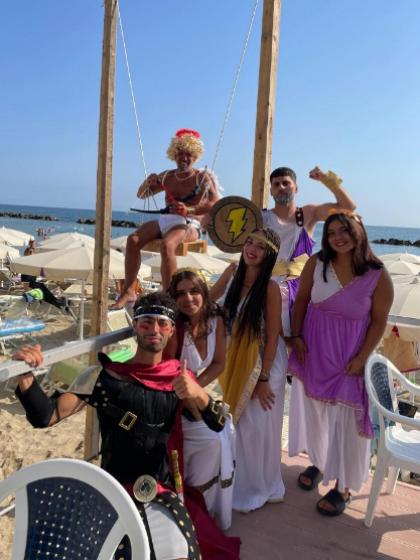 Persone in costumi romani e greci posano su una spiaggia con sfondo marino.