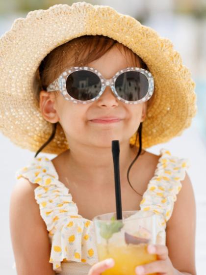 Bambina con cappello di paglia e occhiali, beve una bevanda rinfrescante.