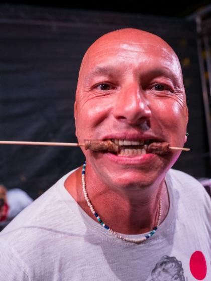 Uomo sorridente con spiedino di carne in bocca, atmosfera festosa.